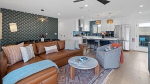 A modern living room with a brown leather sofa, a grey armchair, and a coffee table. A kitchen with white cabinets and a stainless steel refrigerator is visible in the background. - Walk S Congress Pool Putt Putt 3 KING/2 BATH FULLY Remodeled (Austin)