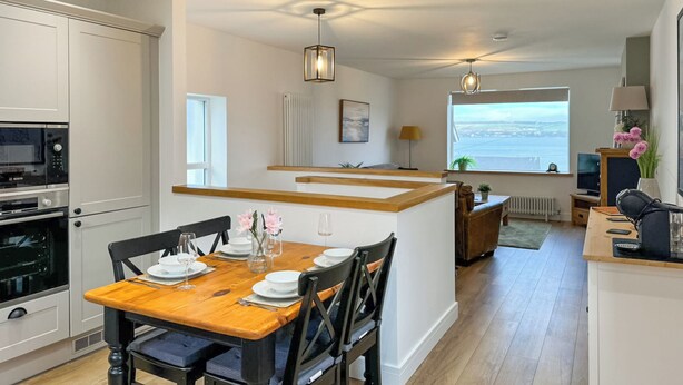 Een moderne keuken met witte kasten, een houten eettafel en uitzicht op zee door een groot raam.