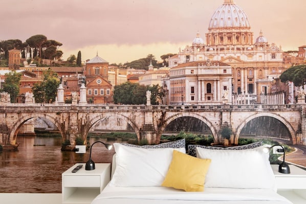 A bedroom with a bed, white pillows, a yellow pillow, a nightstand with a lamp, and a mural of a historic cityscape with a bridge and buildings.