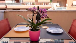 Una mesa con una maceta rosa, dos platos y un vaso en un balcón con sillas rojas y un edificio al fondo.