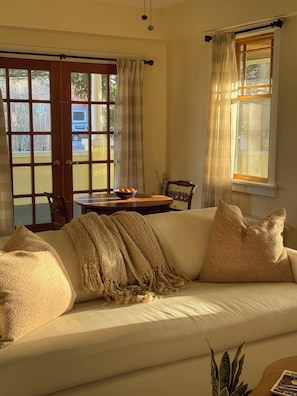 A cozy living room with a sofa, a table with a bowl of fruit, and a view of the outside through large windows.