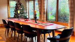 Un comedor con una mesa de madera, sillas negras, un árbol de Navidad y ventanas grandes con cortinas.