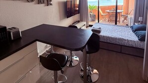 Una habitación de hotel moderna con kitchenette, un comedor con dos taburetes tipo bar, un sofá y un balcón con vista al mar.