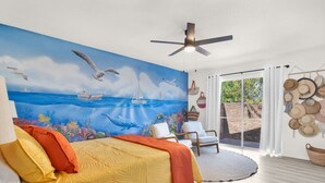 Un dormitorio con cama, ventilador de techo, un mural del océano, ventana con vistas al exterior y un muro decorado con sombreros.