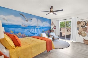 Un dormitorio con cama, ventilador de techo, un mural del océano, ventana con vistas al exterior y un muro decorado con sombreros.