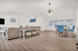 Una habitación amplia con suelo de madera, un sofá blanco, una mesa de comedor con sillas y un ventanal grande.