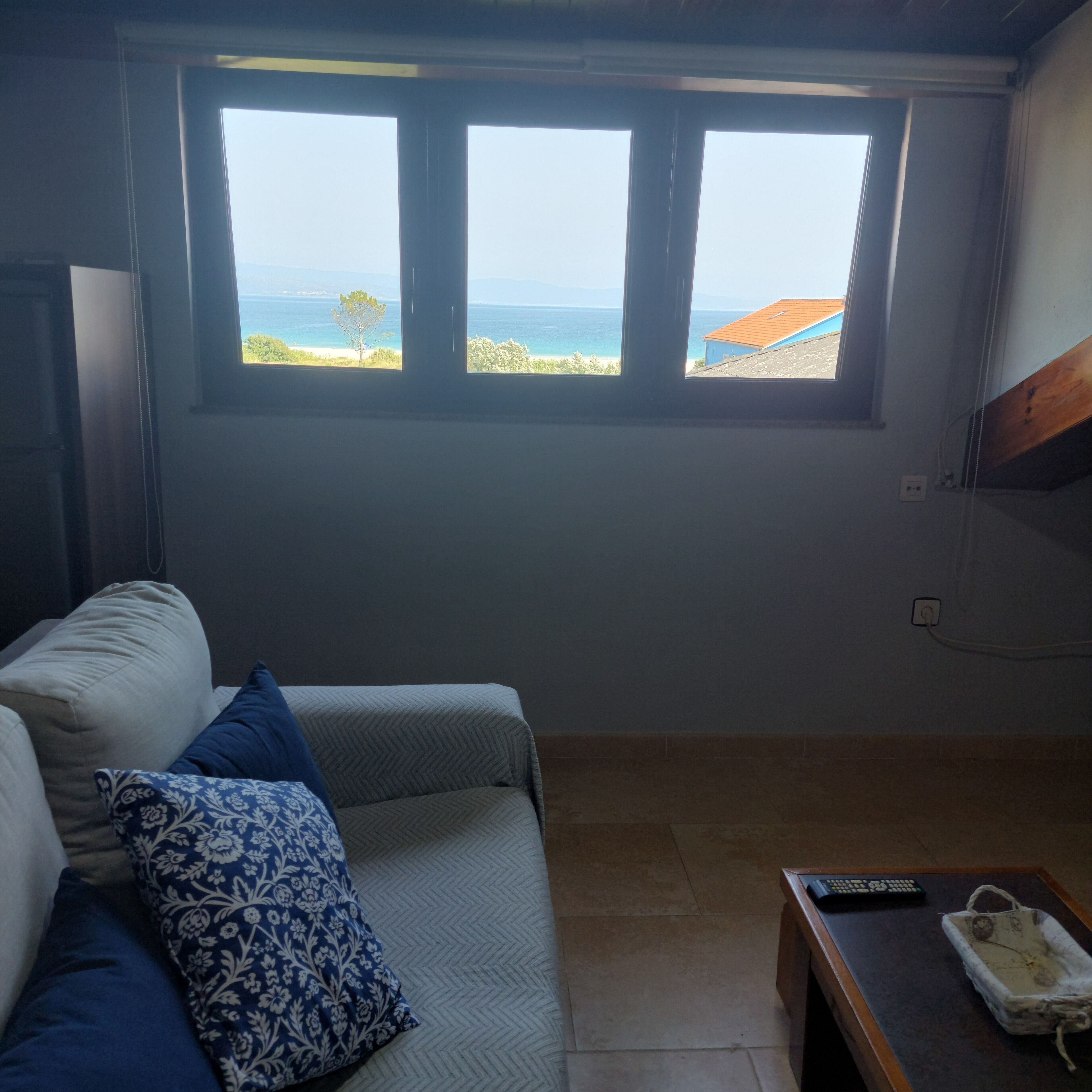 A room with a view of the sea through a window, a sofa with patterned cushions, and a small table with a remote control.