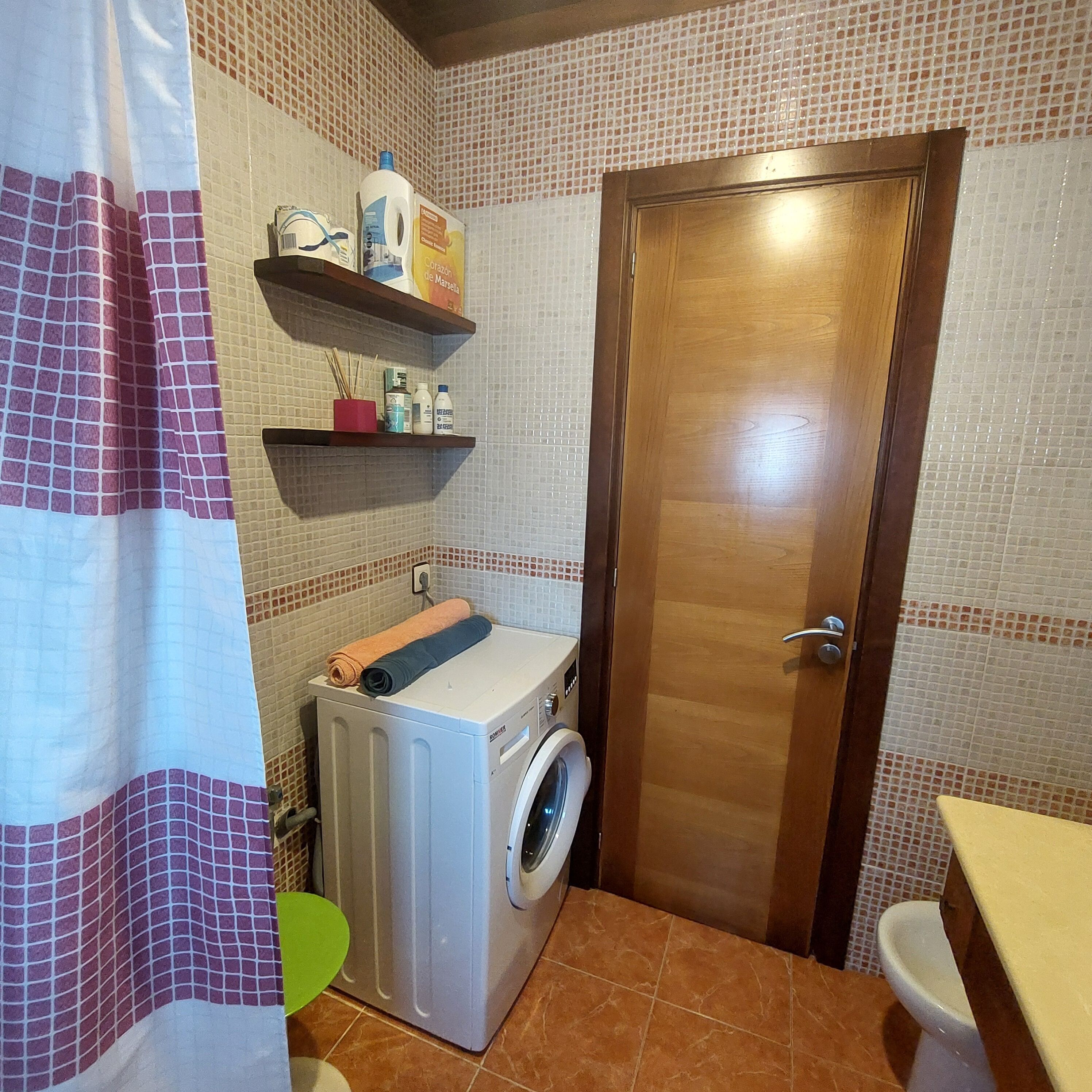 Une petite salle de bain équipée d’un lave-linge, de toilettes et d’une étagère où sont rangés les articles de toilette.