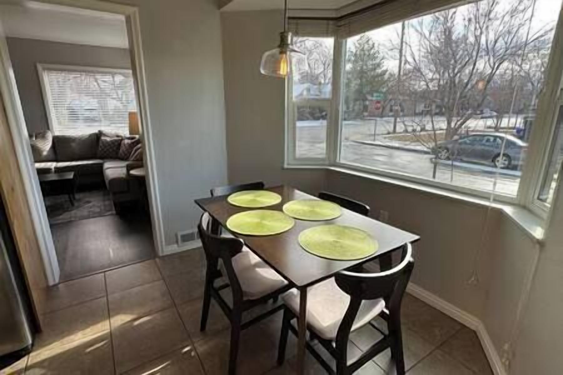 A dining area with a table set for four, a bay window with a view of the street, and a sofa in the adjacent living room.