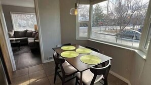 A dining area with a table set for four, a bay window with a view of the street, and a sofa in the adjacent living room.