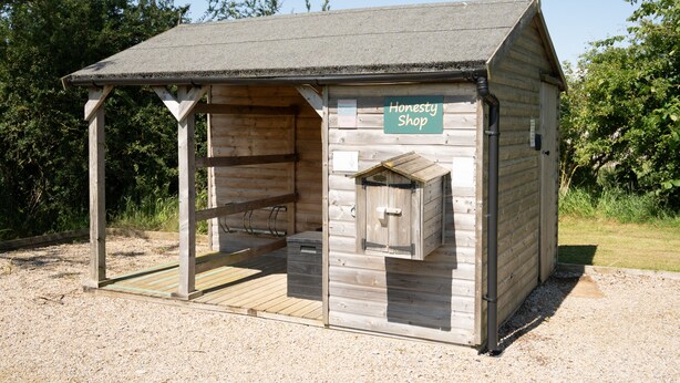Una struttura in legno con un tetto spiovente, un piccolo portico e una porta in legno, con la scritta 'Honesty Shop'.