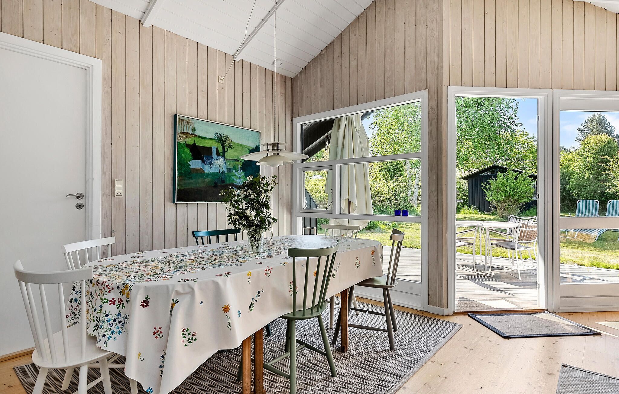 A dining area with a table set for four, a painting on the wall, and a view of the garden through large windows.