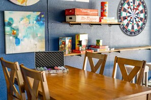 A dining area with a wooden table and chairs, a dartboard, shelves with snacks, and a painting on the wall. - Pool House w/ Outdoor Bar, Game Room & Movie Room (Irving)