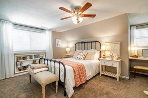 A bedroom with a bed, bedside tables, a desk, a chair, a ceiling fan, and a window with blinds.