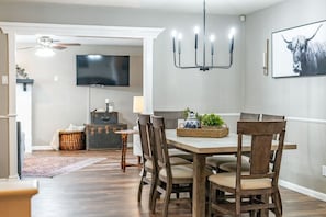 A dining area with a wooden table and chairs, a chandelier, and a mounted TV. - Pool House w/ Outdoor Bar, Game Room & Movie Room (Irving)