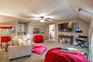 A cozy attic bedroom with a ceiling fan, a red bean bag chair, a white sofa, and a desk with a TV.