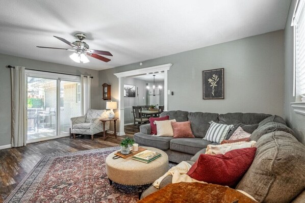 A living room with a sectional sofa, a round ottoman, a coffee table, and a patterned rug. There is a ceiling fan, a dining area in the background, and a sliding glass door leading to a patio.