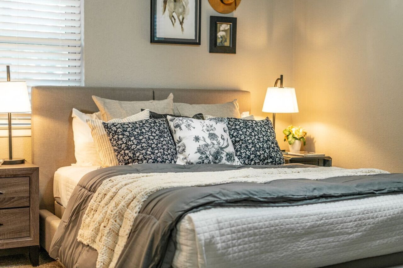 A neatly made bed with floral-patterned pillows, a bedside lamp, and a small plant on the nightstand.