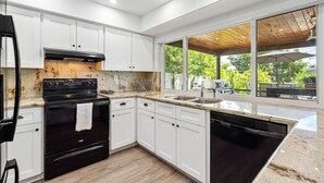 A modern kitchen with white cabinets, a black stove, and a large window overlooking a patio area. - BBQs & Breezes: Waterfront Pool House Escape (Margate)