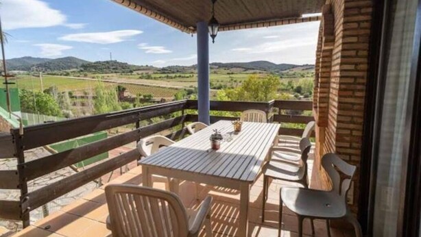 A covered outdoor patio with a table and chairs, overlooking a rural landscape with fields and hills.