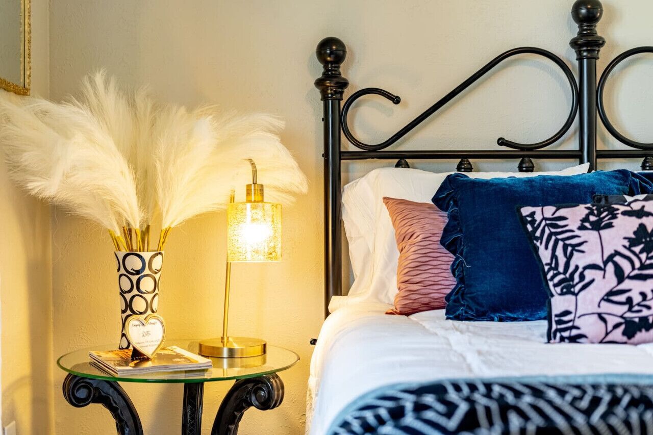 A neatly made bed with a patterned bedspread, a decorative vase with feathers, and a bedside lamp on a glass table.