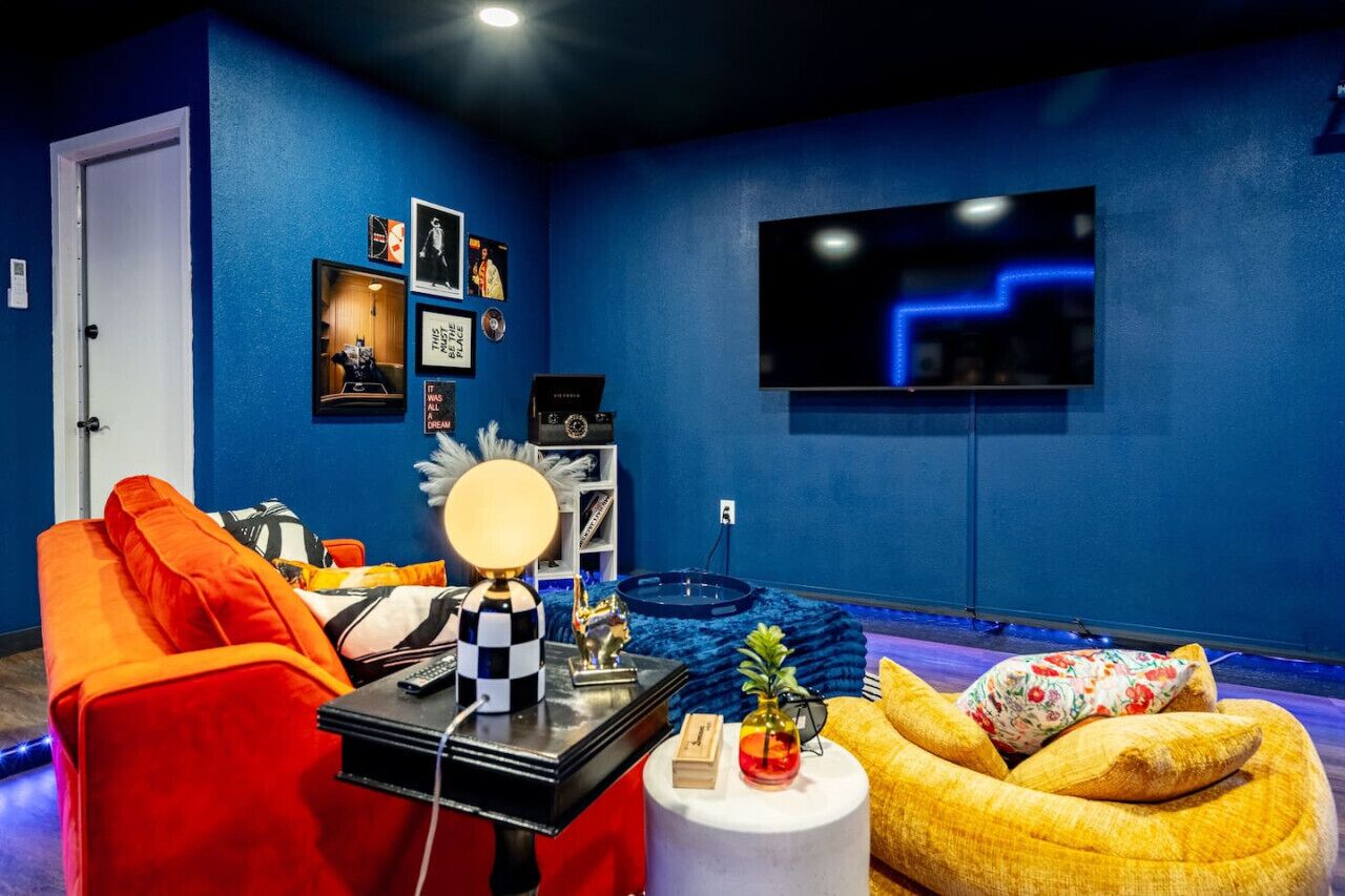 A room with a blue wall, a large flat-screen TV, a red sofa, a yellow bean bag, and a small round table with a lamp.