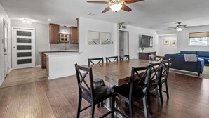 A modern kitchen with wooden cabinets, a dining area with a wooden table and chairs, and a living room with a blue sofa. - Tropical Pool w/ Tiki Bar, Fire Pit, Swing (Richardson)