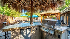 An outdoor grill area with a thatched roof, a pool, and lounge chairs. - Tropical Pool w/ Tiki Bar, Fire Pit, Swing (Richardson)
