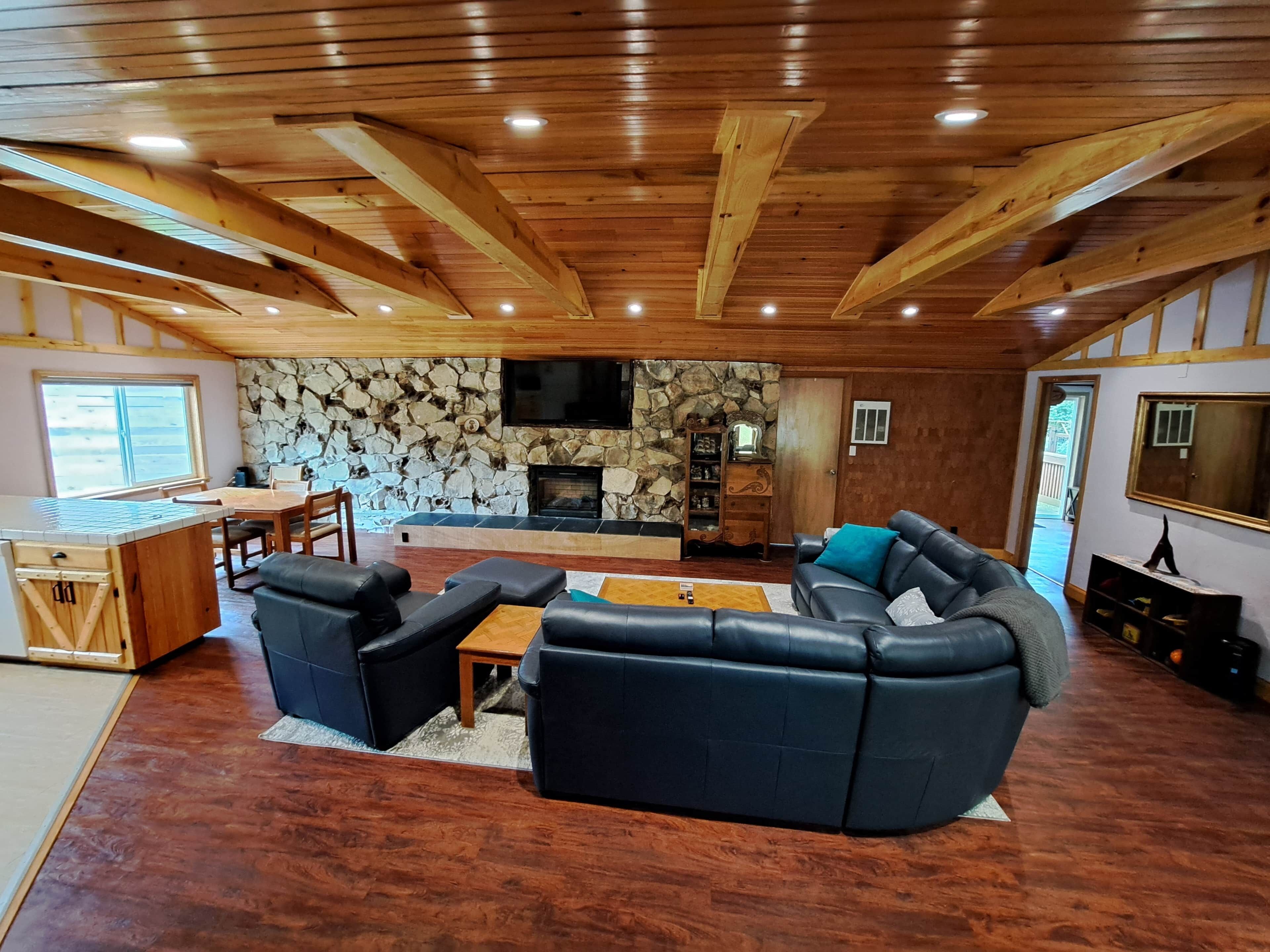Un vaste espace de vie agrémenté d’une cheminée en pierre, de poutres apparentes en bois et d’un grand canapé en cuir noir.