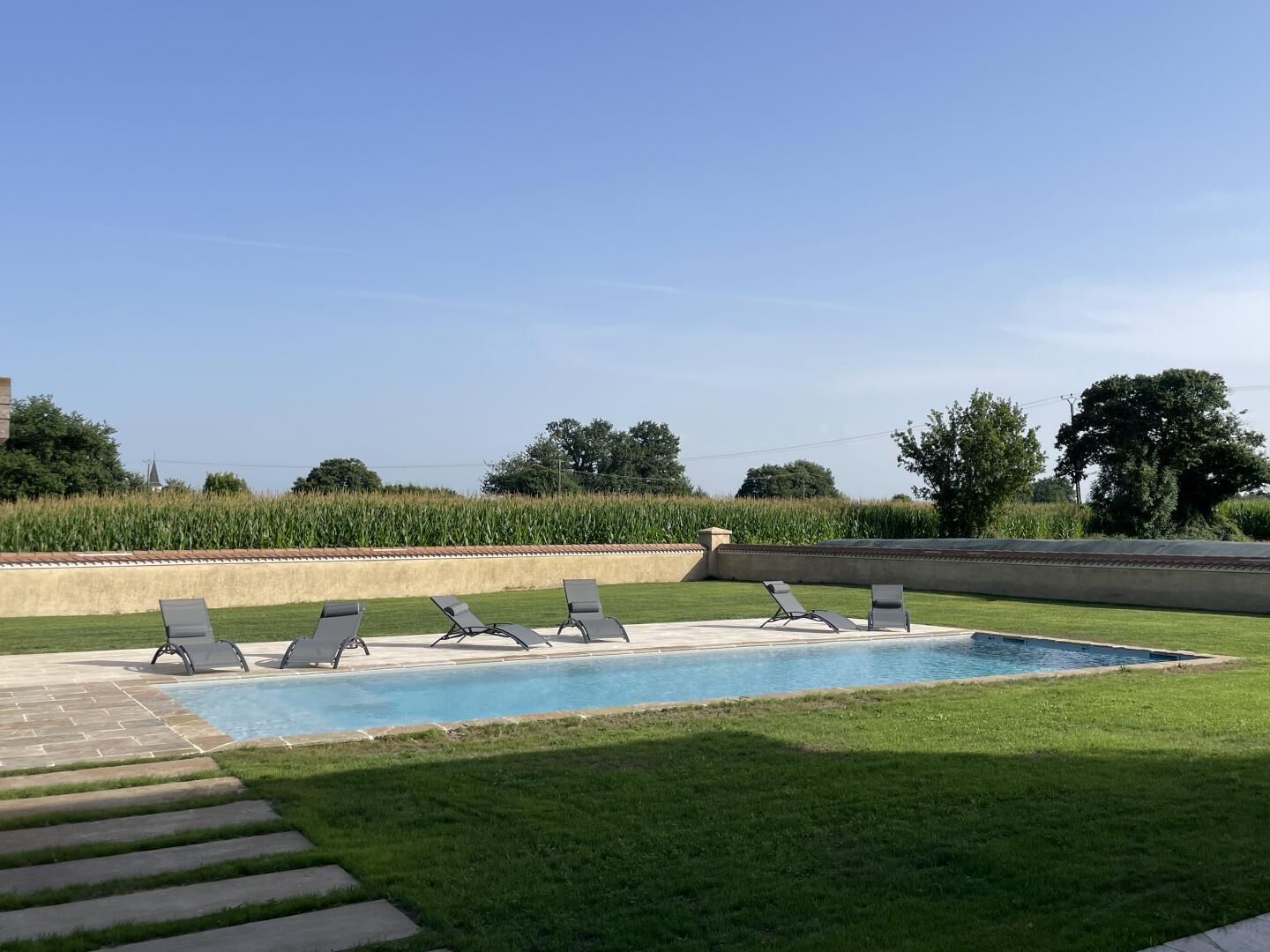 Una piscina con sillones de descanso, un espacio con césped y vistas a un campo de maíz y árboles.