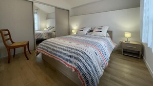 A bedroom with a large bed, a wooden chair, a bedside table with a lamp, and a mirror on the wall. - TOUTOU - ANGLET - Gîte (ANGLET)