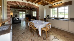 Une cuisine rustique avec une cheminée en briques, des poutres apparentes en bois au plafond et un coin repas avec une table dressée pour quatre personnes.