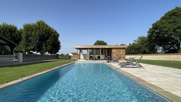 Une piscine rectangulaire à l’eau d’un bleu limpide, entourée d’une terrasse carrelée. On aperçoit, au fond, des chaises longues et une pergola en bois.