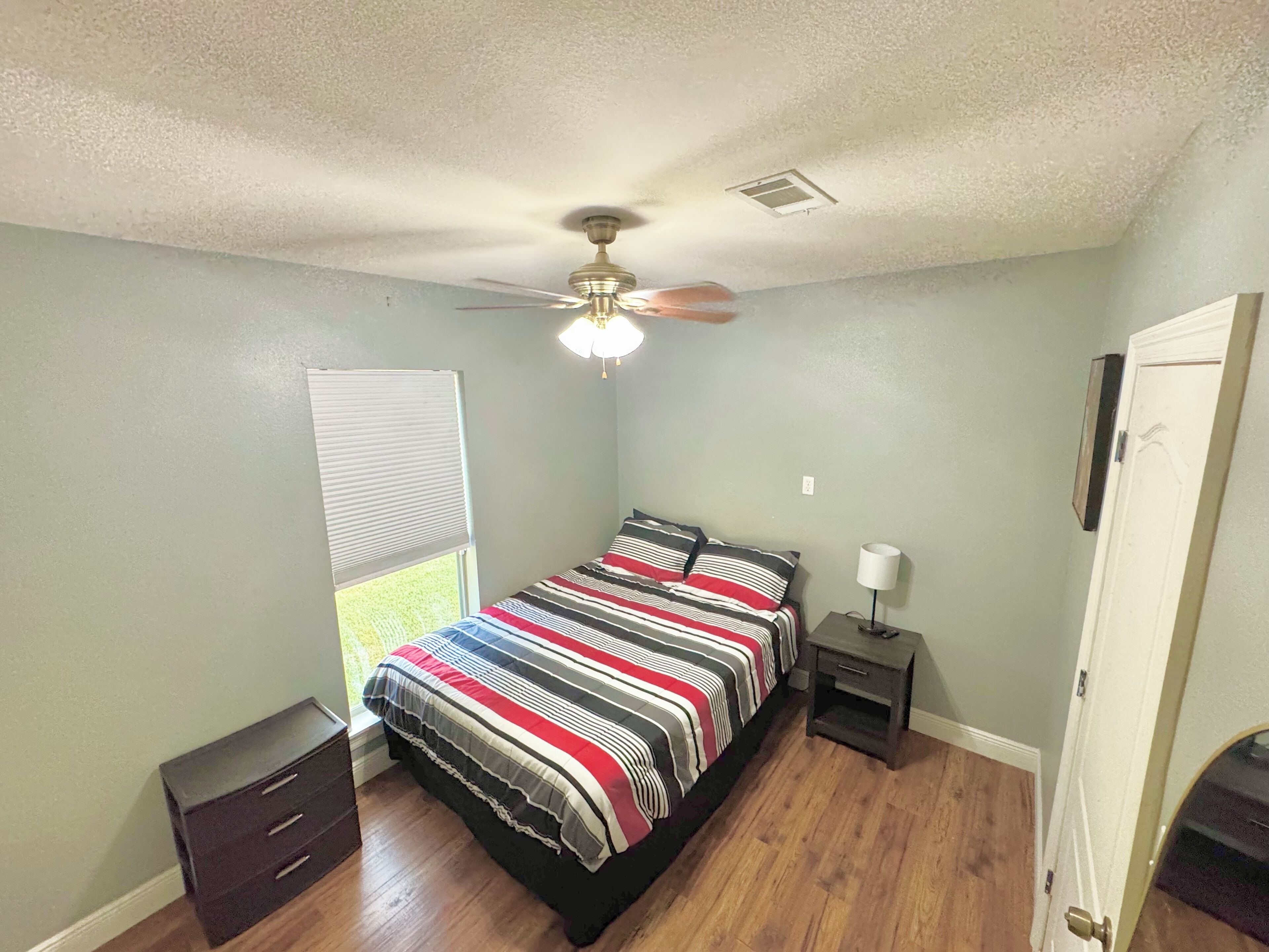Un dormitorio con cama, ventilador de techo, mesita de noche, ventana con persianas y puerta.