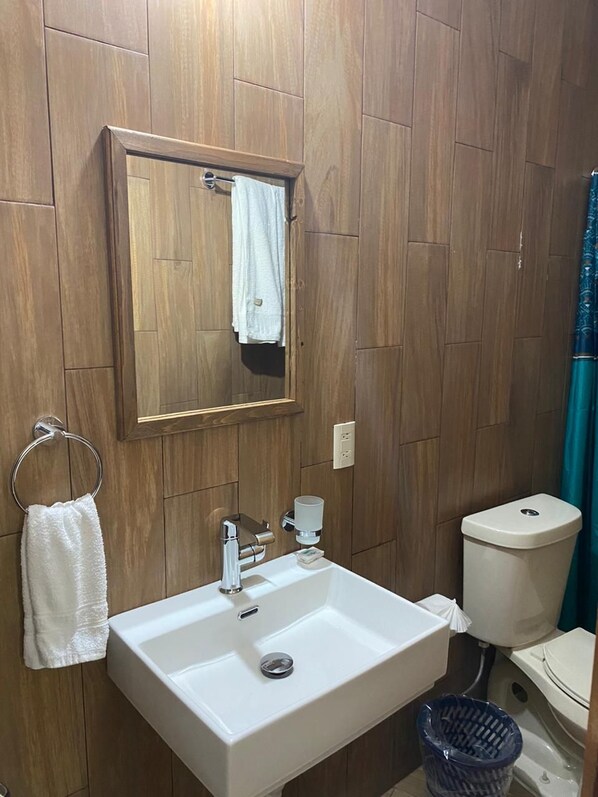 A bathroom with wooden paneling, a white sink with a mirror above it, a towel rack, and a blue trash bin. - Posada Conchita (Tlalpujahua)