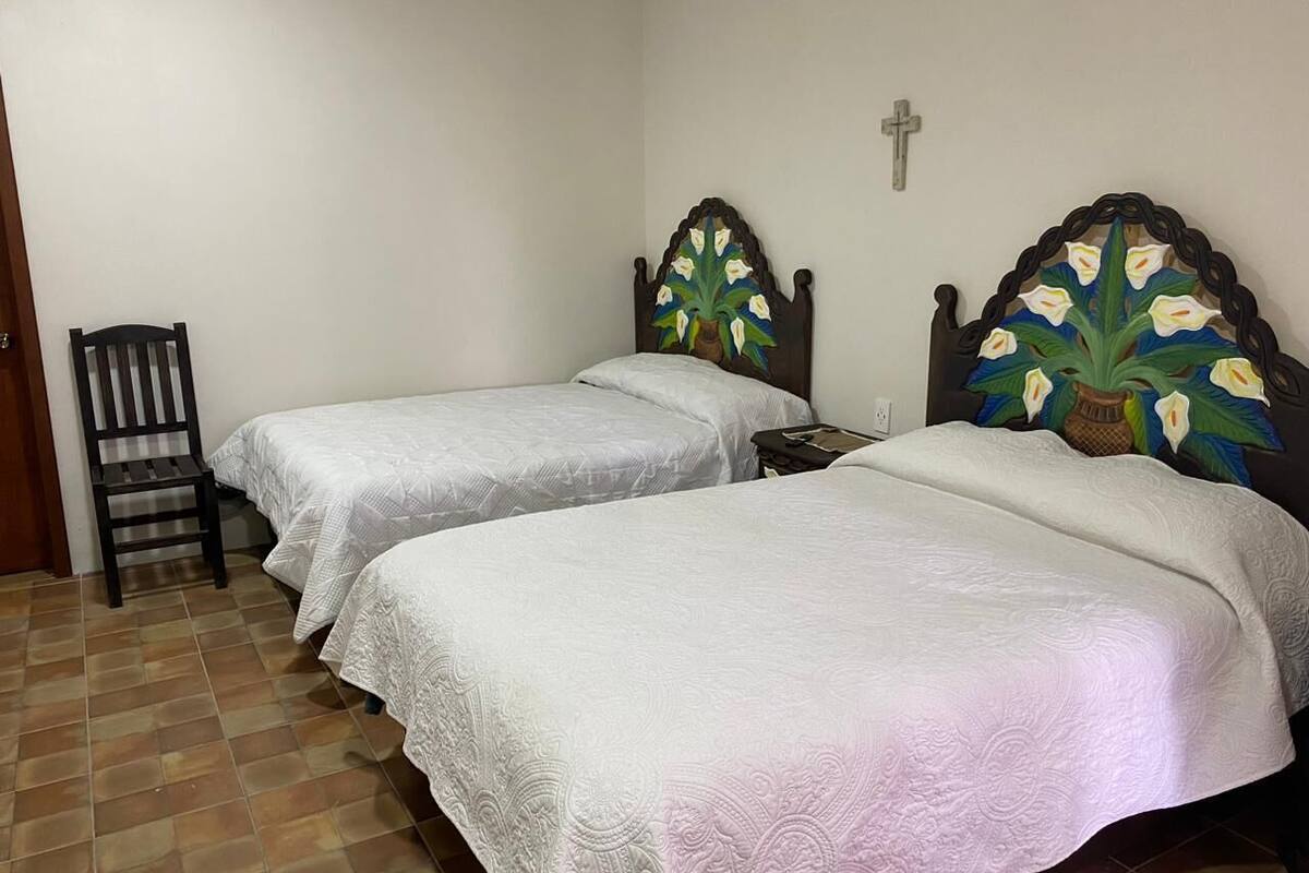 A room with two single beds, a wooden chair, and a wall with a cross and a decorative headboard.
