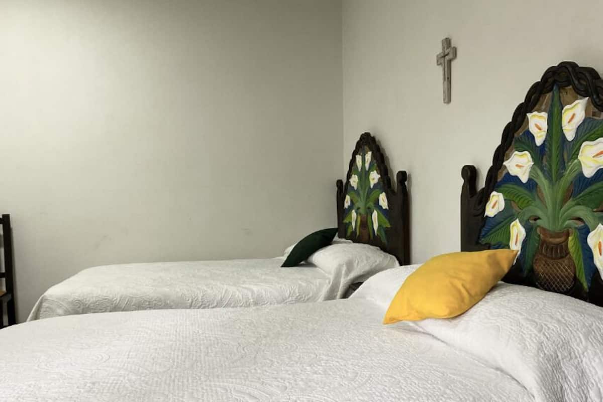A room with two beds, each with white bedding and a single pillow. There is a wooden chair and a wall-mounted cross.