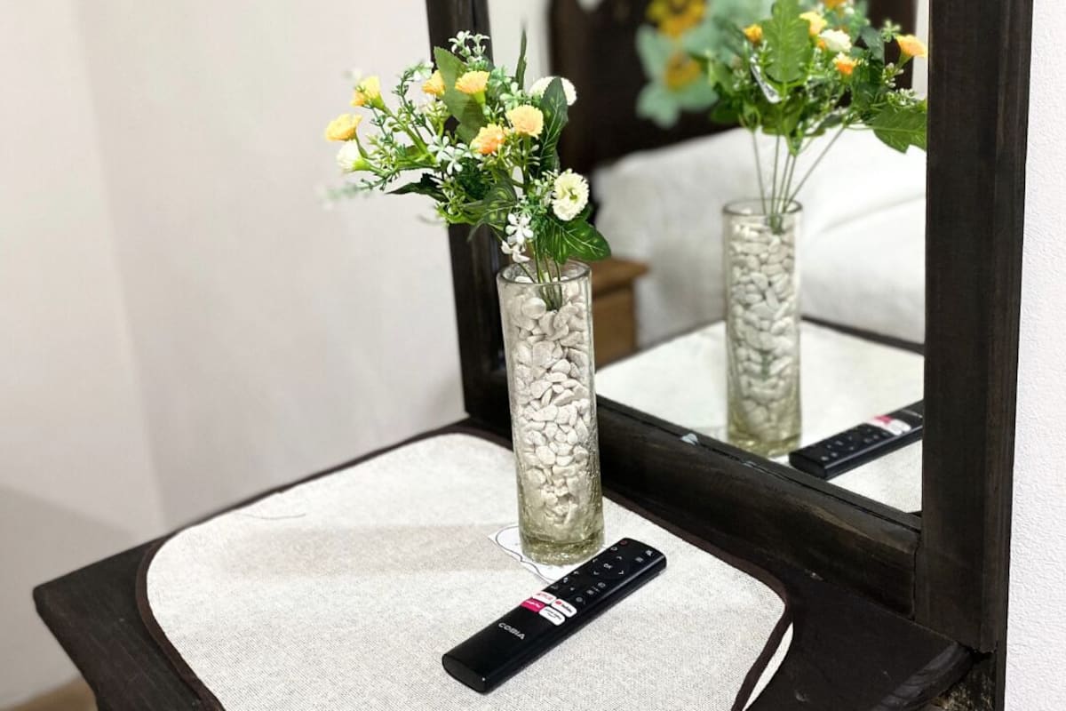 A mirror reflecting a bed with a floral headboard, a remote control on a textured mat, and a vase with flowers on a bedside table.