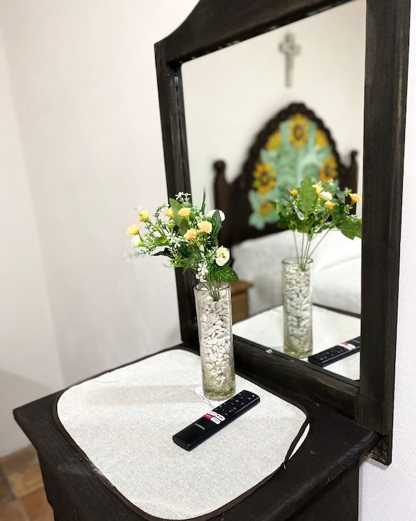 Um espelho reflete uma cama com cabeceira florida, um controle remoto sobre um tapete texturizado e um vaso com flores em uma mesinha de cabeceira.