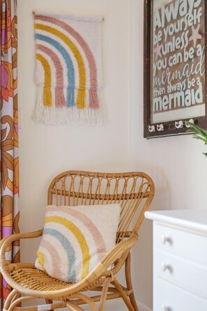 A wicker chair with a rainbow-patterned pillow, a wall with a mermaid-themed quote, and a patterned curtain. - Hygge By The Sea | Beachfront & Pet Friendly (Avon)