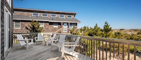 A wooden deck with white chairs, a building with stone facade, and a clear view of the beach.