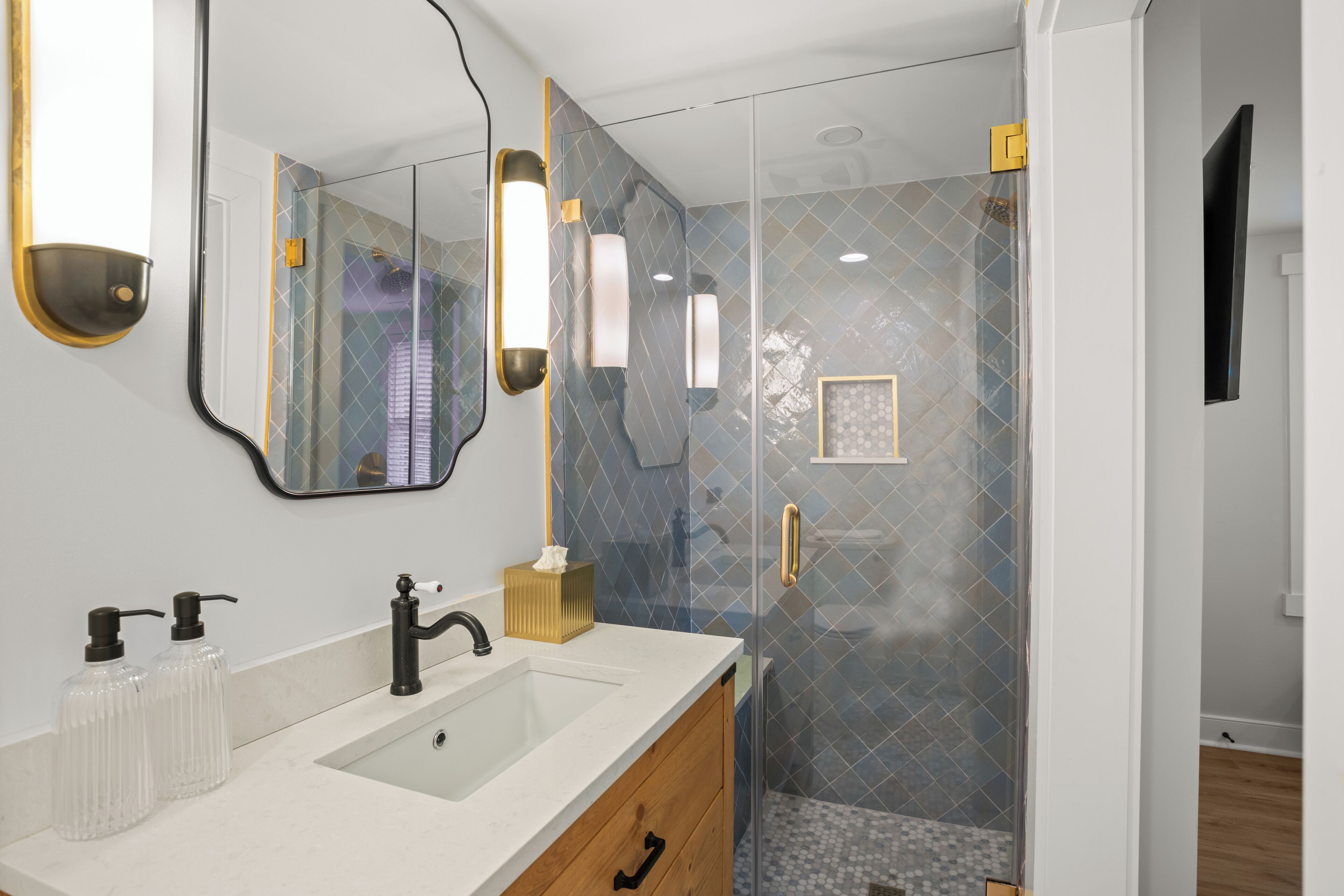 Un baño moderno con ducha de vidrio, lavamanos blanco y espejo con marco dorado.