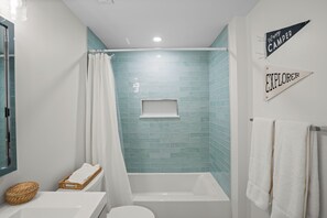 Un baño con una bañera blanca, un inodoro blanco y una zona de ducha con pared de azulejos azules. Hay dos toallas colgadas en la pared y una cesta sobre el mostrador.