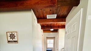 Un couloir étroit avec un plancher en bois, des murs blancs et un plafond en bois. Une poubelle est visible sur la gauche, et une porte se trouve sur la droite.