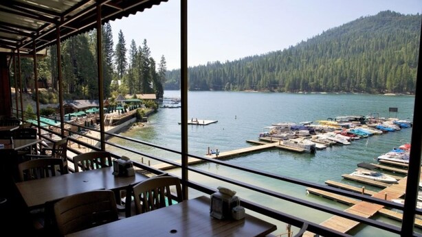 A lakeside restaurant with outdoor seating overlooking a docked boat area and a forested hill.