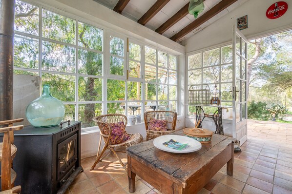 A sunlit room with large windows, wicker chairs, a wooden table, and a vintage stove.