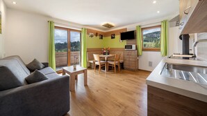 Una sala moderna con sofá, mesa de comedor y sillas, una cocina con pileta y un televisor de pantalla plana, y ventanas amplias con cortinas.