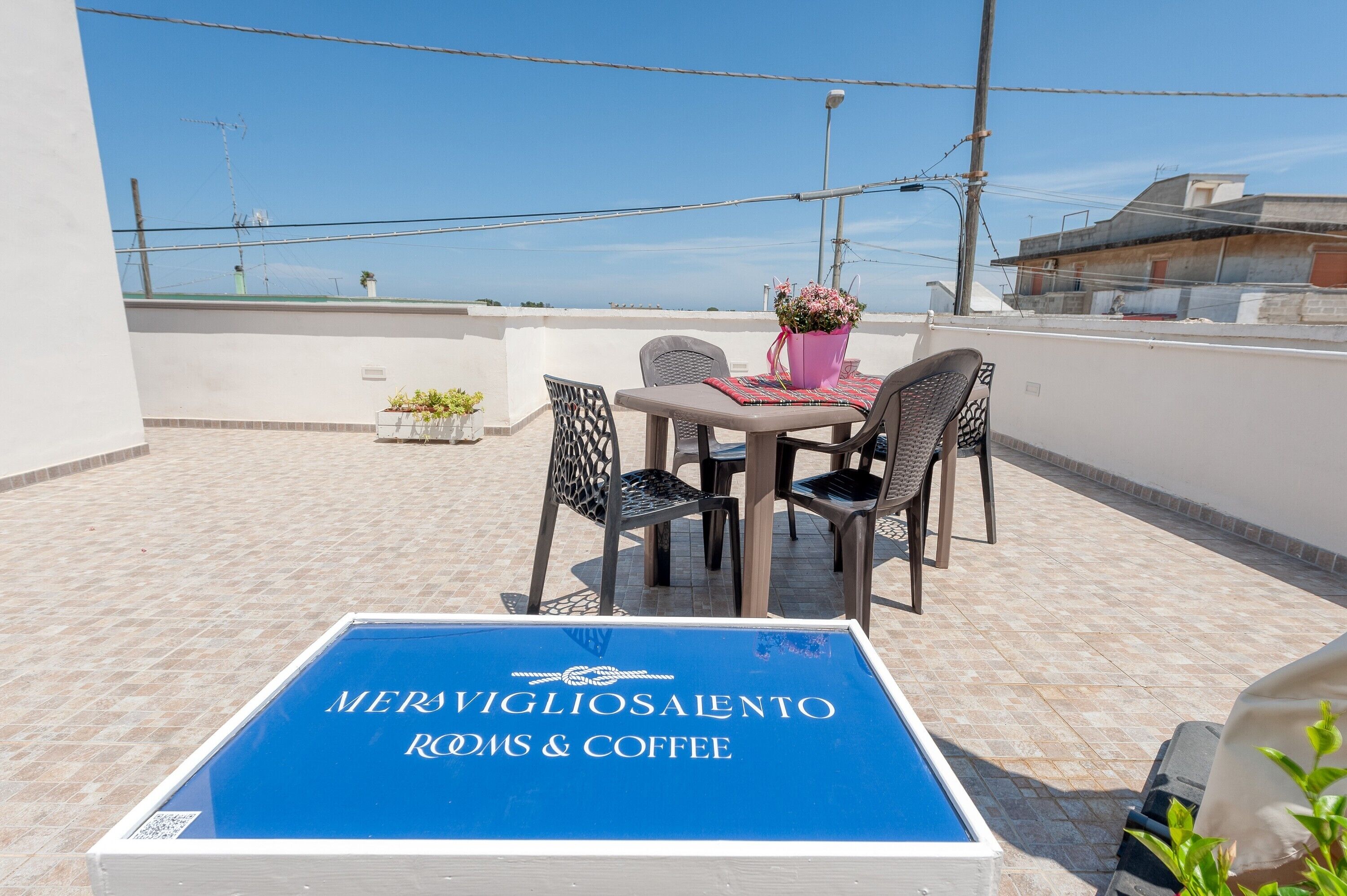 A rooftop terrace with a table and chairs, a flower vase, and a sign for 'MERS VIGLIOSALENTO ROOMS & COFFEE'.