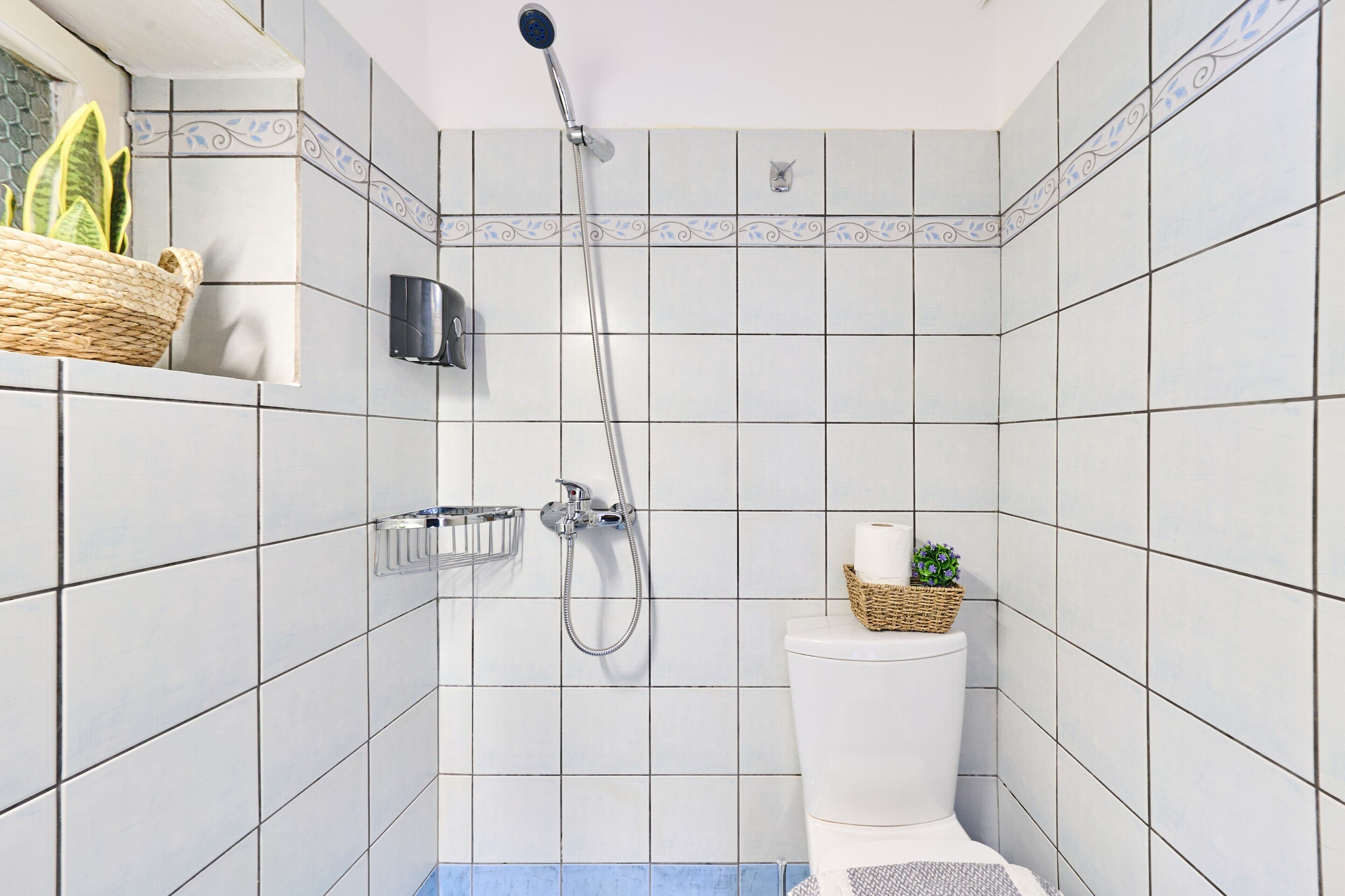 Ein Badezimmer mit weißen Fliesen, einer Dusche, einer Toilette, einem Seifenspender und einer kleinen Pflanze.