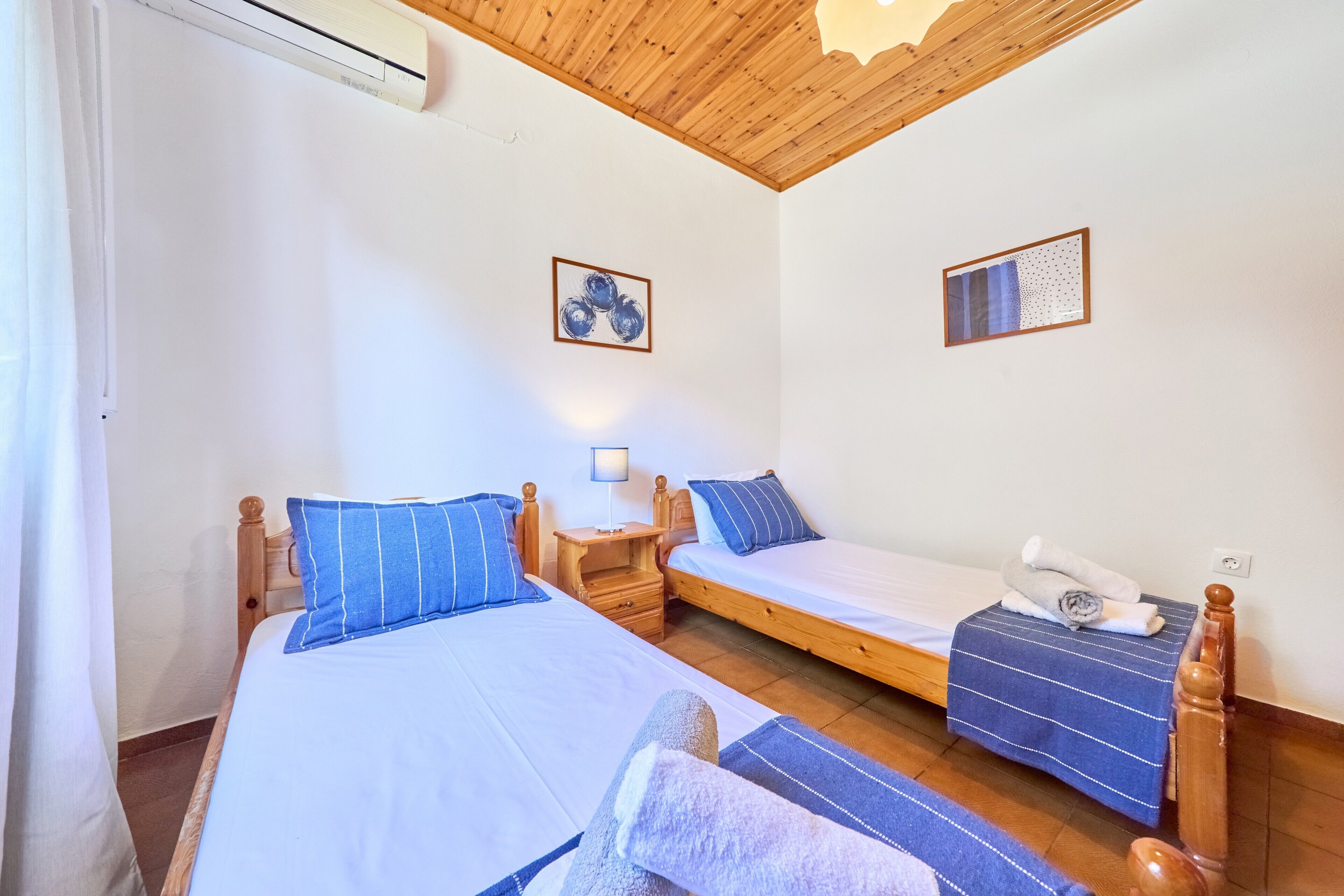Een kamer met twee bedden, houten hoofdeinden, een nachtkastje en een wandairconditioner.